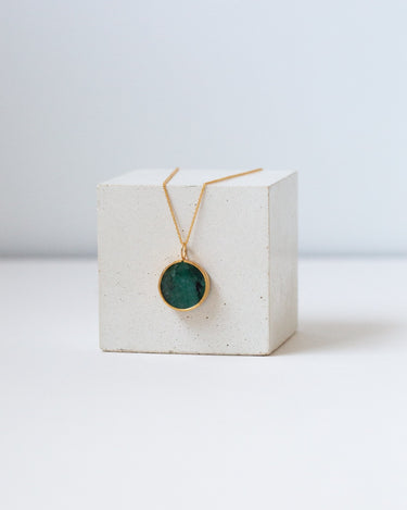 Round emerald pendant on a gold chain draped over the white cube.