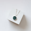 Round emerald pendant on a silver chain draped over the white cube.