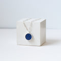 Round lapis lazuli pendant on a silver chain draped over a white cube.