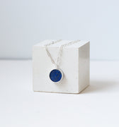Round lapis lazuli pendant on a silver chain draped over a white cube.
