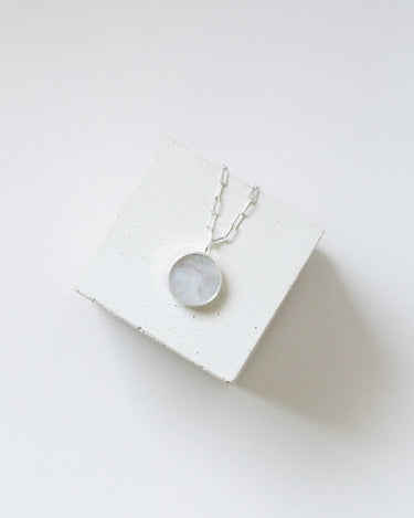 Round moonstone pendant on a silver chain draped over a white cube.